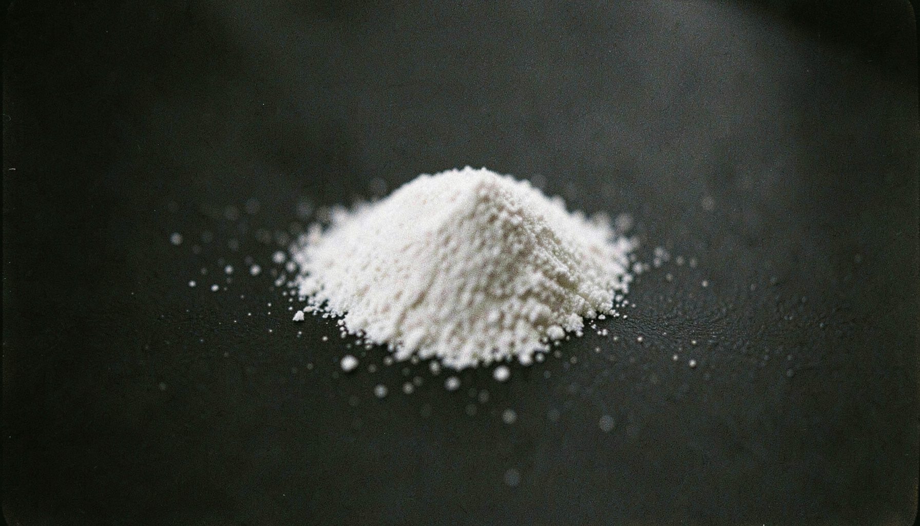 A close-up analog film photograph with visible grain of a fine white crystalline powder resting on a dark matte surface, moody and subdued lighting with deep charcoal shadows.