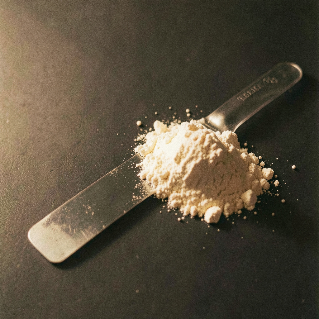 A laboratory spatula beside a small pile of off-white powder on a dark surface, side-lit to emphasize texture, warm amber and charcoal tones.