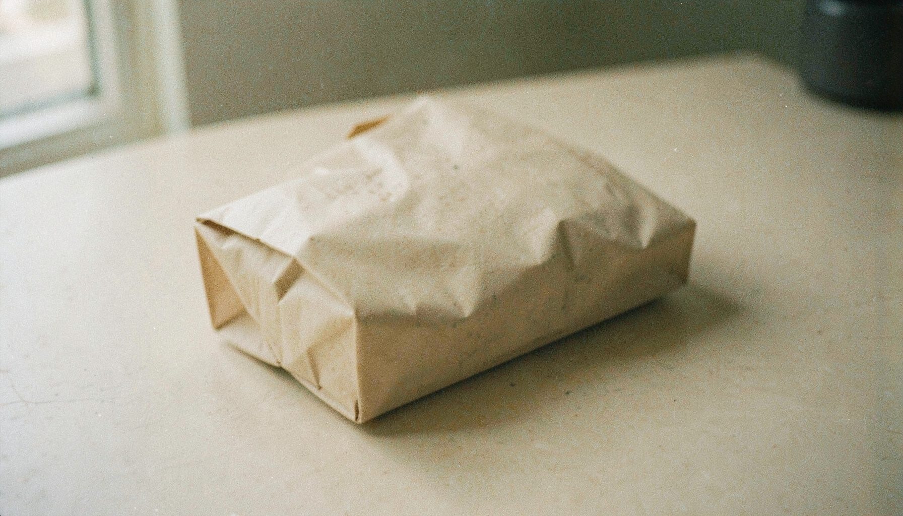 Film photograph of a sealed discreet shipping package on a plain table, neutral beige tones, soft natural window light with visible film grain.