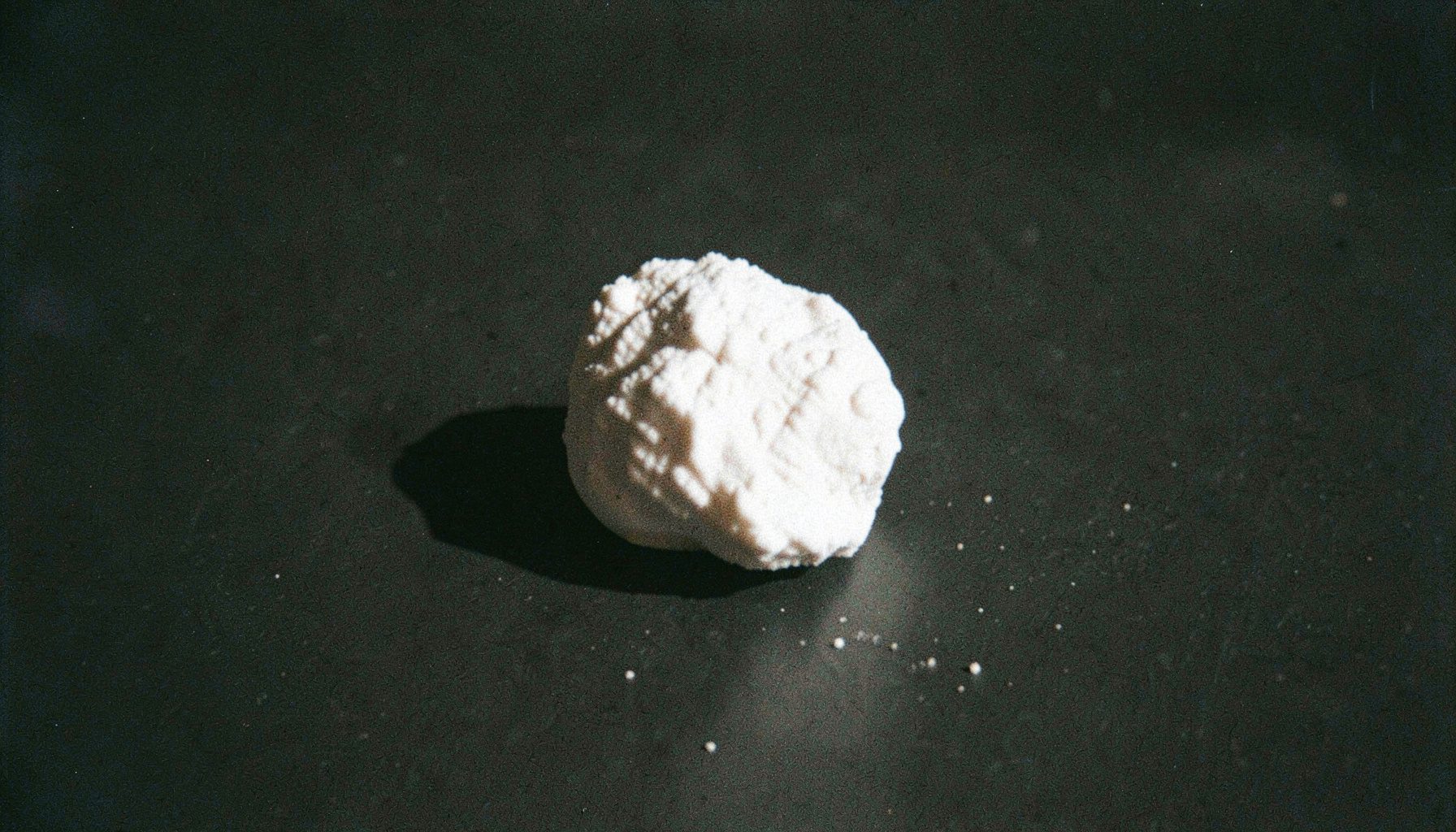 Grainy analog photograph of white powder formed into a small rock-like chunk on a dark matte surface, dramatic side lighting casting long shadows.
