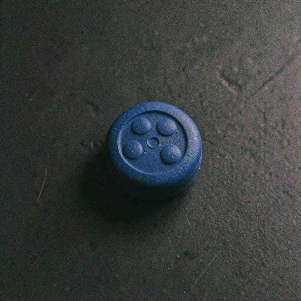A grainy film photograph of a deep blue pill with a domino dot pattern embossed on its face, placed on a dark matte surface with subtle, moody lighting.
