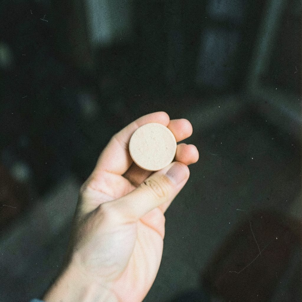 A grainy film photograph of a hand holding a small round tablet between thumb and forefinger against a dark, out-of-focus background, with soft natural light and visible film grain.