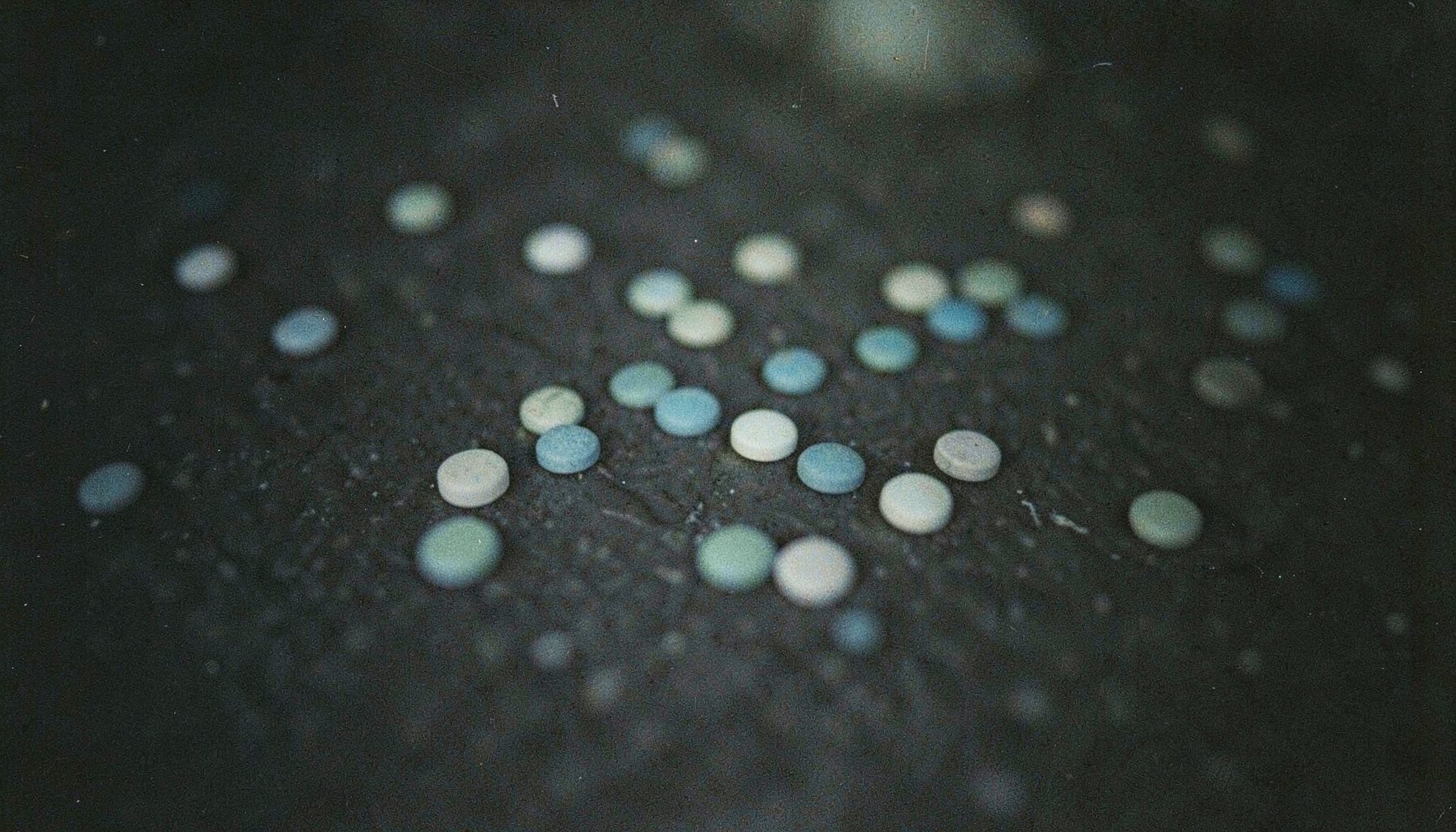 A dark, moody close-up photograph with visible film grain showing scattered small round tablets in muted blues, greens, and off-whites on a textured dark surface, with soft shadows and a subdued, clandestine atmosphere.