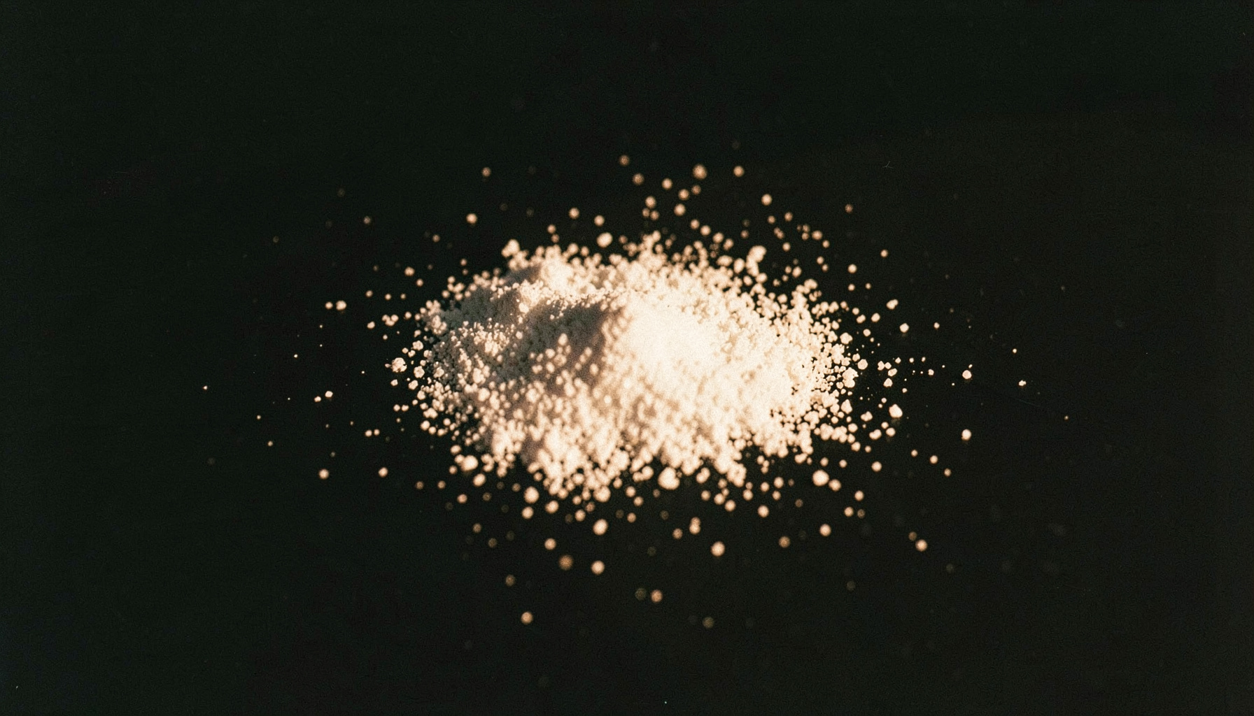 A dark, moody close-up photograph of scattered white crystalline powder on a matte black surface, lit from one side with a single warm tungsten light casting long shadows. The grain is visible and the overall atmosphere is stark and clandestine.